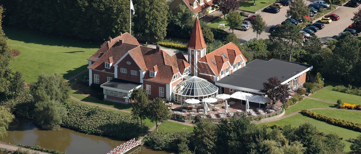 Sørup Herregård set fra luften — rødstensbygning med tårn og glasoverdækket havestue, sø og park omkring